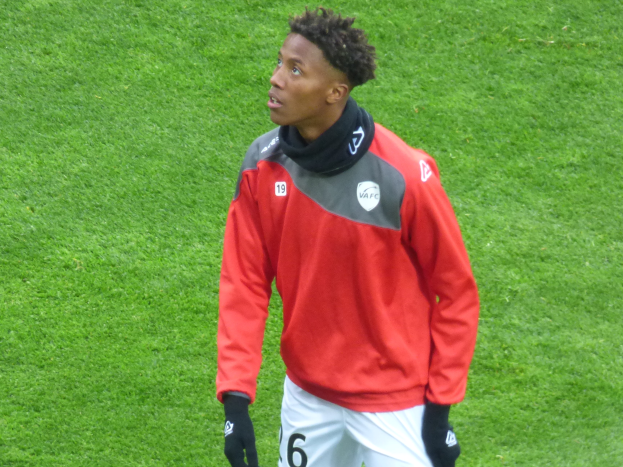 Ein junger Mann in einer roten Jacke und weißen Shorts steht auf einem Fussballfeld, schaut nach rechts mit einem entschlossenen Gesichtsausdruck, trägt Baseball-Handschuhe.