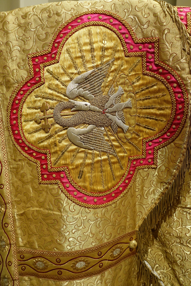 Nahaufnahme eines Priestergewandes mit goldener und roter Stickerei vor einer Wand im Hintergrund, mit detaillierter und luxuriöser Verarbeitung.