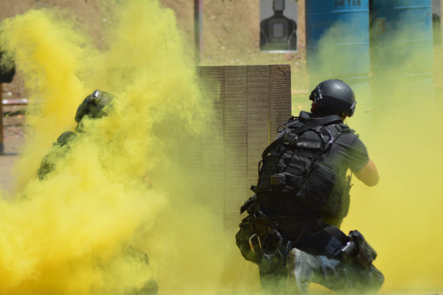 Zwei Polizisten in Einsatzkleidung mit gelbem Rauch aus dem Mund, stehend in der Nähe einer Wand mit Text und anderen Gegenständen im Hintergrund.