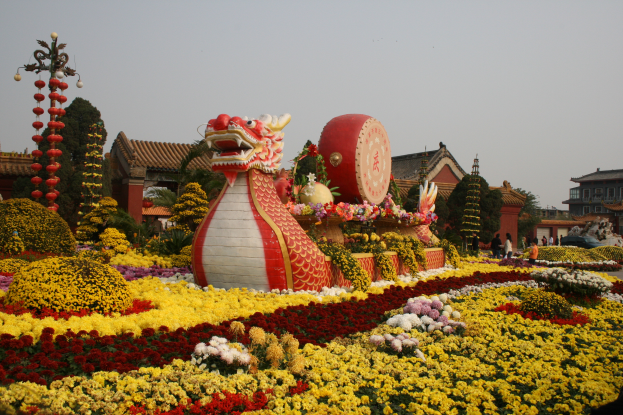 Ein chinesisches Neujahrsfest mit einer Drachenstatue, umgeben von farbenfrohen Blumen, mit Menschen, Gebäuden, Bäumen und einem klaren blauen Himmel im Hintergrund.