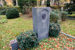 Ein Friedhof mit einem zentralen Grabstein, umgeben von Bäumen, Pflanzen und Gras, mit Gebäuden im Hintergrund.