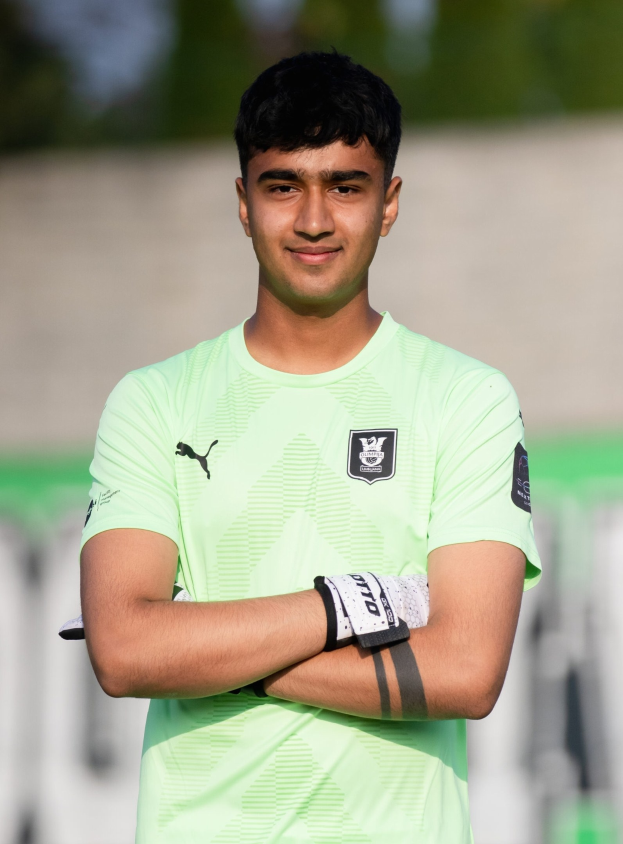 Ein junger Mann in grünem T-Shirt steht auf einem Fußballfeld mit verschräckten Armen, lächelnd, mit einem unscharfen Hintergrund.
