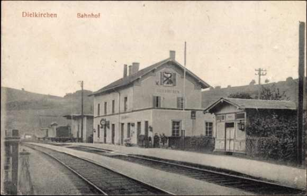 Schwarz-weiß-Fotografie eines Bahnhofs in Dielkirchen, Deutschland, mit einem Zug auf den Schienen, umgeben von Häusern, Strommästen, Bäumen, Menschen und Hügeln im Hintergrund unter einem klaren Himmel.