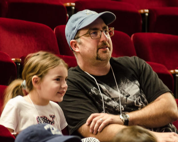 Ein Mann mit Brille, Uhr und Mütze sitzt mit einem strahlenden kleinen Mädchen in einem Kino, im Hintergrund sind Stühle und andere Menschen zu sehen.