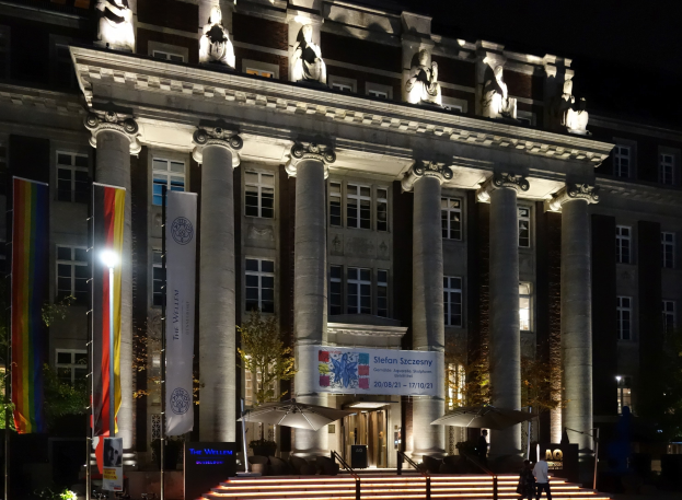 Neoklassisches Gebäude mit hohen Säulen und Statuen auf seinem Dach, nachts beleuchtet, umgeben von Bäumen, Fahnen, Bannern, Treppen, Geländern und Schirmen, mit einigen Menschen und einem dunklen Himmel im Hintergrund, identifiziert als das Willem Dafoe Museum in Berlin, Deutschland.