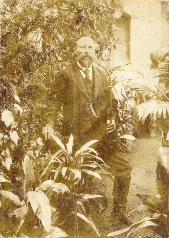 Altes Schwarz-Weiß-Foto eines Mannes in Anzug und Krawatte, der in einem Garten mit Pflanzen und Bäumen steht, eine Wand im Hintergrund sichtbar.