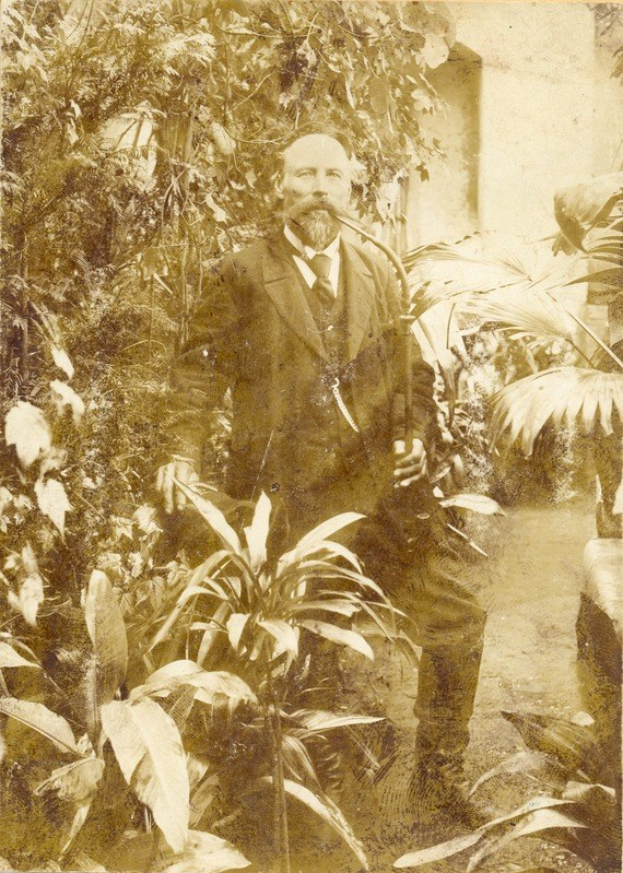 Altes Schwarz-Weiß-Foto eines Mannes in Anzug und Krawatte, der in einem Garten mit Pflanzen und Bäumen steht, eine Wand im Hintergrund sichtbar.