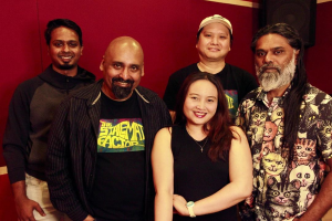 Eine Gruppe von Menschen in legerer Kleidung, die eng zusammenstehen, lächeln und vor einer roten Wand mit einem Lautsprecher im Hintergrund stehen, was darauf hindeutet, dass sie sich in einem Aufnahmestudio befinden.