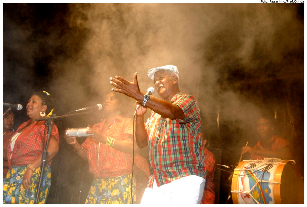 Eine Gruppe von Musikern, die auf der Bühne stehen, wobei ein Mann in der Mitte in ein Mikrofon singt und ein anderer Mann rechts ein Schlagzeug spielt.