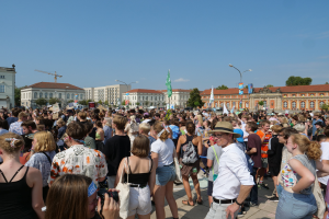 Eine große Menschenmenge steht vor einem Gebäude mit Fenstern, Laternenmasten, Flaggen und Bäumen, einige halten Kameras, was auf eine Protest- oder Demonstrationsveranstaltung unter einem bewölkten Himmel hindeutet.