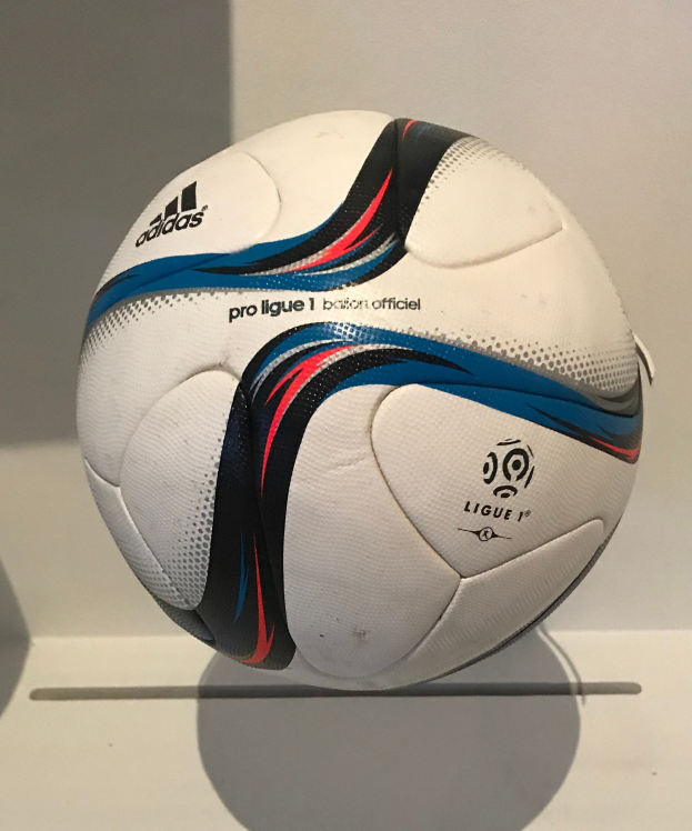 Ein weißer Fußball mit schwarzem Adidas-Logo und 'Ligue 1'-Aufdruck liegt auf einem weißen Tisch vor einem hellen weißen Hintergrund.