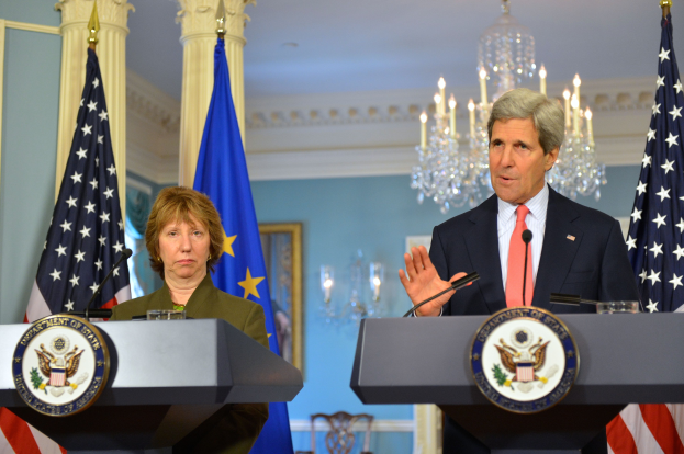 Außenminister John Kerry und der Minister für Innere Sicherheit stehen an Podien im East Room des Weißen Hauses, mit Kerry, der in ein Mikrofon spricht, zwei Flaggen dahinter und einer Kristallleuchte im Hintergrund.