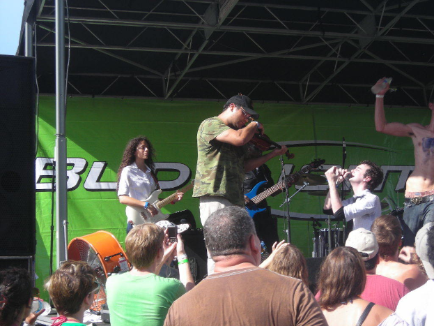 Eine Gruppe Musiker spielt auf der Bühne mit verschiedenen Instrumenten, während das Publikum vor einem Podium steht und die Musik genießt. Ein großer schwarzer Lautsprecher ist links positioniert, und ein grünes Banner ist im Hintergrund sichtbar.