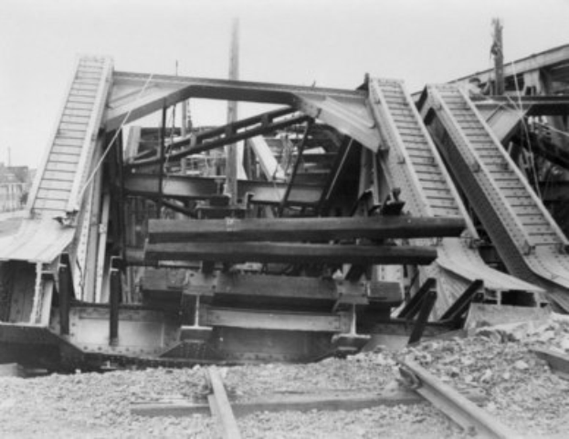 Schwarz-weiß-Foto einer Bahntrasse, die zu einer zerfallenden Brücke mit kaputten Geländern und Schutt führt, umgeben von Gebäuden unter einem klaren Himmel.
