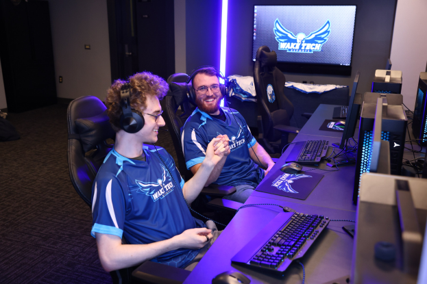 Zwei Männer in blauen T-Shirts und Kopfhörern sitzen an einem Schreibtisch mit Computerausrüstung und spielen ein Spiel in einem Raum mit einem Bildschirm, Lampen und Taschen auf dem Boden.