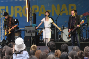 Eine Bühne mit vier Musikern: eine Frau in einem weißen Kleid in der Mitte, zwei Männer mit Gitarren links und rechts daneben und eine Menge und Lautsprecher im Hintergrund.