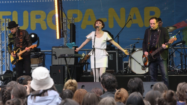 Eine Bühne mit vier Musikern: eine Frau in einem weißen Kleid in der Mitte, zwei Männer mit Gitarren links und rechts daneben und eine Menge und Lautsprecher im Hintergrund.
