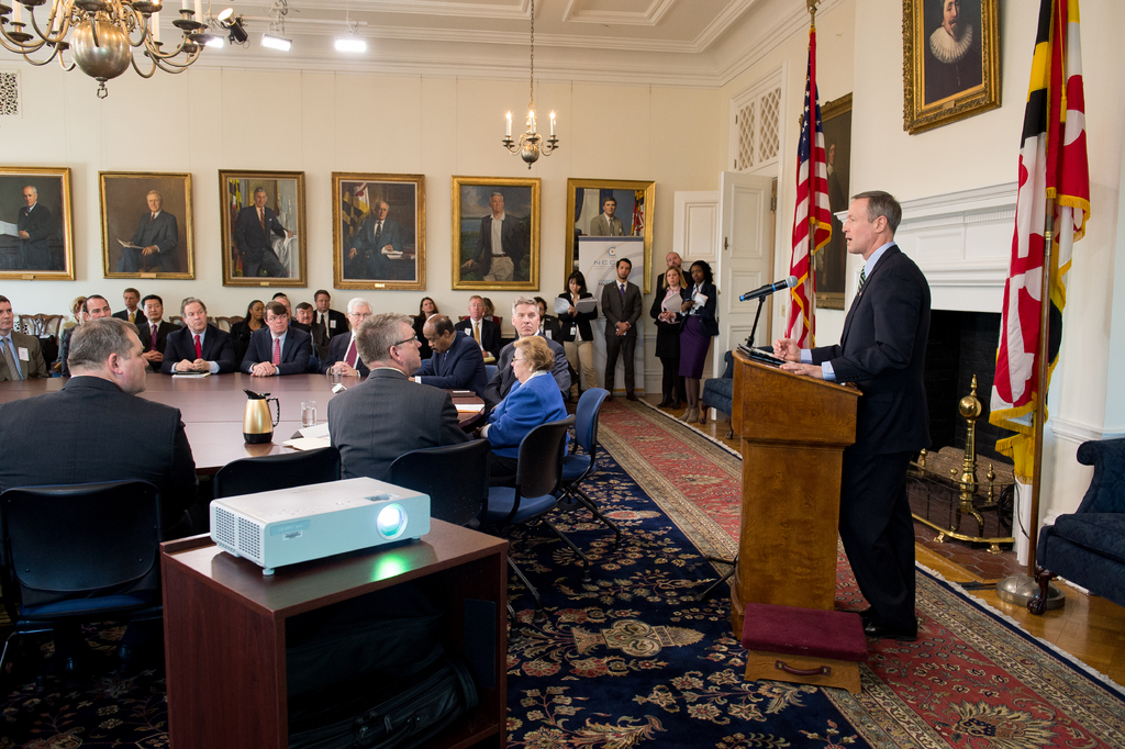 Ein Mann steht rechts vor einem Rednerpult und spricht in ein Mikrofon, während einige Personen links auf Stühlen sitzen und ihm zuhören. Auf dem Tisch links steht ein Projektor. An der Wand hängen Foto Rahmen. Rechts sind zwei Flaggen zu sehen. Oben im Bild sind Lampen und eine Deckenleuchte zu erkennen.