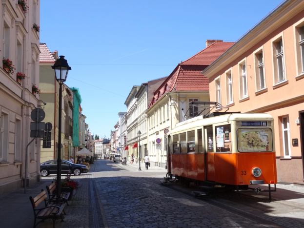 Eine orangefarbene Straßenbahn fährt durch die kopfsteingepflasterte Straße der Altstadt von Tallinn, flankiert von Gebäuden mit Fenstern, Bänken und Laternen, mit Menschen, die spazieren gehen, und dem Himmel im Hintergrund.