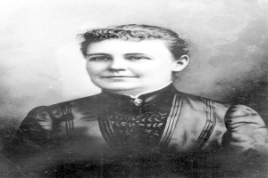 Schwarzes und weißes Foto einer Frau in einem Kleid, die mit verschränkten Händen und erhobenem Kopf steht und Entschlossenheit und Stärke ausstrahlt.