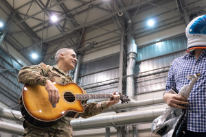 Ein Mann in Militäruniform spielt eine Gitarre, während er neben einem anderen Mann in einem Helm steht, mit Rohren und Lichtern im Hintergrund.