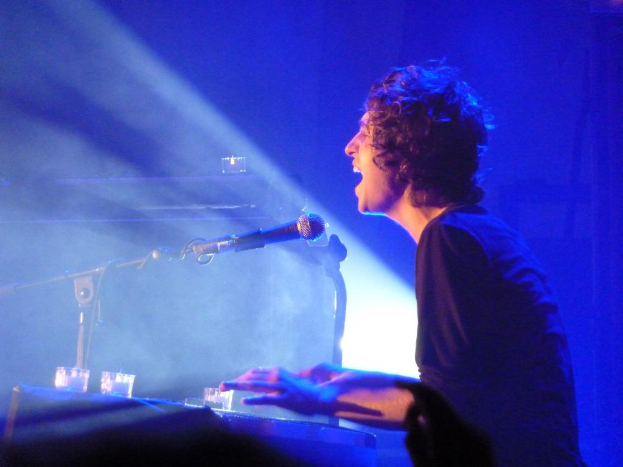 Eine Person in schwarzem T-Shirt ist als DJ mit einem Mikrofon vor ihr zu sehen, vor blauem Hintergrund.