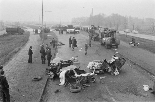 Schwarz-weiß-Szene eines Autounfalls am Straßenrand mit mehreren Fahrzeugen, Menschen drumherum, Laternen, Bäume und Himmel im Hintergrund.
