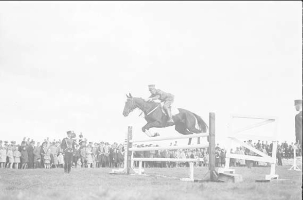 Ein Schwarz-Weiß-Foto eines rotbraunen Pferdes mit einer weißen Blesse, schwarzer Mähne und Schweif, das über ein hölzernes Hindernis springt, während ein Reiter in einem weißen Hemd und blauen Jeans zusieht, mit einer Zuschauermenge im Hintergrund.