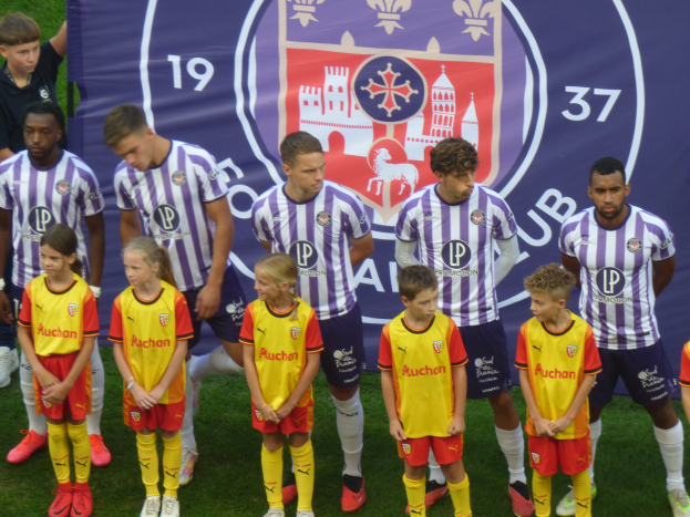 Eine Gruppe junger Menschen, die auf einem Fußballfeld stehen und ein Banner mit dem Text 'Preston Primary School Football Team' und Bildern im Hintergrund halten.