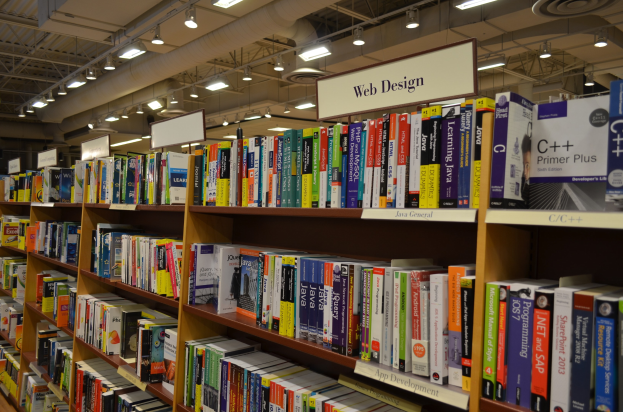 Ein Bibliotheksinneres mit Bücherregalen, Informationsplakaten, Deckenleuchten und sichtbaren Rohren.