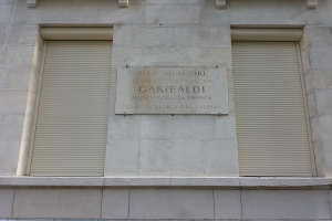 Ein zweistöckiges Gebäude mit zwei Fenstern und Rollläden sowie einer Gedenktafel an der Außenwand.