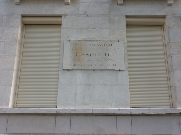 Ein zweistöckiges Gebäude mit zwei Fenstern und Rollläden sowie einer Gedenktafel an der Außenwand.