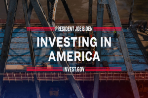 Präsident Joe Biden in der Nähe einer Brücke mit dem Text "In America investieren" im Vordergrund und verstreuten Gegenständen auf dem Boden.