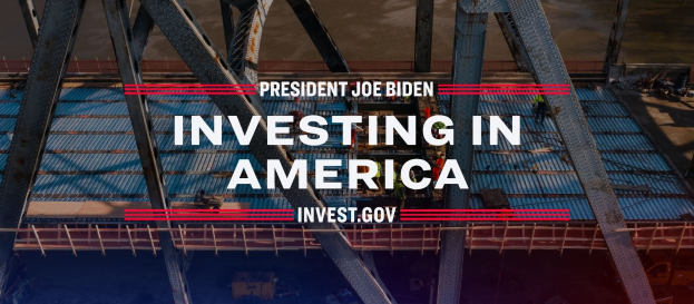 Präsident Joe Biden in der Nähe einer Brücke mit dem Text "In America investieren" im Vordergrund und verstreuten Gegenständen auf dem Boden.