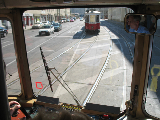 Ein Mann fährt eine Straßenbahn auf einer Stadtstraße mit anderen Fahrzeugen, Gebäuden und einem klaren blauen Himmel im Hintergrund.