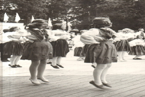 Schwarz-weisses Foto von Frauen in traditioneller bayerischer Tracht, die auf einem Holzfussboden tanzen, umgeben von Baelmen und Fahnen im Hintergrund.