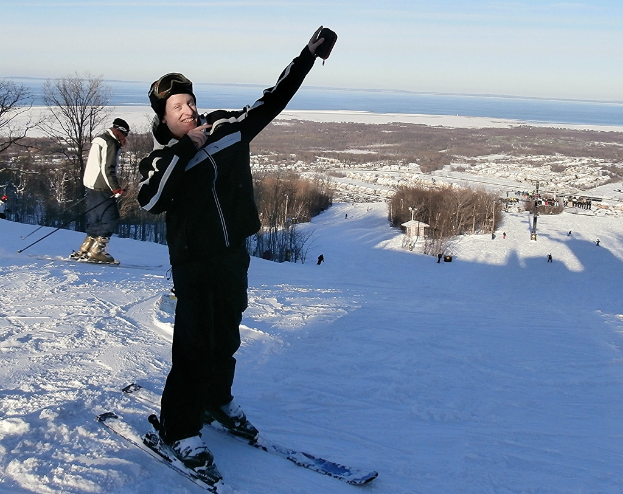Eine Person mit einem Snowboard steht zentral auf einer verschneiten Landschaft, im Hintergrund sind Skifahrer, Bäume, Menschen, ein Pfahl, Wasser und der Himmel zu sehen.