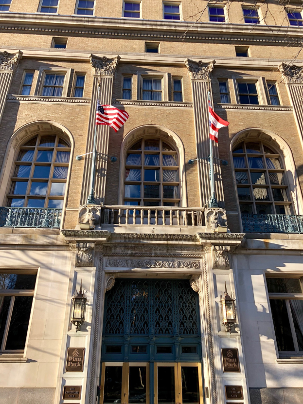 Außenansicht des Plaza Hotels in New York City, mit Glasfenstern, Türen, Säulen, Geländern, Leuchten, Texttafeln und Flaggen vor einem Himmel-Hintergrund.