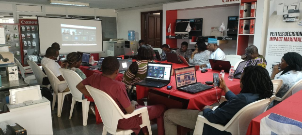 Gruppe von Menschen an einem Tisch mit Laptops, Flaschen und anderen Gegenständen während eines digitalen Marketing-Workshops, mit einem Banner, einem Display, einer Tür und Deckenleuchten im Hintergrund.