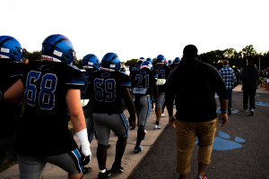 Eine Gruppe von Fussballspielern in Helmen und Stiefeln läuft eine Straße mit Bäumen und einem klaren blauen Himmel im Hintergrund entlang.