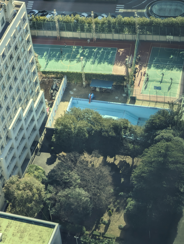 Eine Luftaufnahme eines Tennisplatzes in einer Stadt, umgeben von Gebäuden, Bäumen, Pflanzen, Pfählen, Fahrzeugen auf der Straße und einem Schwimmbad.