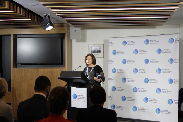 Eine Frau steht auf einer Bühne und spricht in ein Mikrofon, vor ihr sitzt das Publikum, an der Wand hängt ein Banner, ein Fernseher an der Holzwand und Lampen an der Decke.