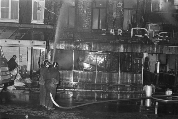 Feuerwehrleute bei der Brandbekämpfung an einer Straßenecke, mit Menschen mit Rohren, einem geparkten Fahrzeug, verstreuten Gegenständen, einem Gebäude mit Fenstern, einem Schild, Drähten und einem bewölkten Himmel.