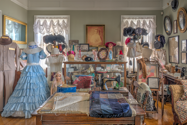 Ein Raum im Museum of Fine Arts in St. Louis, Missouri, voller Schaufensterpuppen in verschiedenen Outfits, ein Tisch mit Kleidung und einer Nähmaschine, Stühle mit Kissen, ein Schrank mit verschiedenen Gegenständen, Fotorahmen an der Wand, Fenster mit Vorhängen und eine Decke.