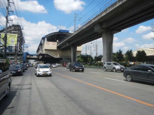 Vollbefahrene Hauptstraße mit fahrenden Autos, eine Brücke im Hintergrund, Strommasten mit Drähten, Bäume, Gebäude und ein bewölkter Himmel.