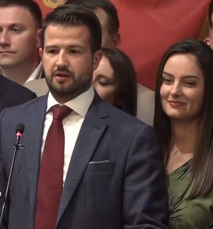 Ein Mann im Anzug steht an einem Rednerpult und spricht in ein Mikrofon, mit einem Banner im Hintergrund und einer Menge davor.