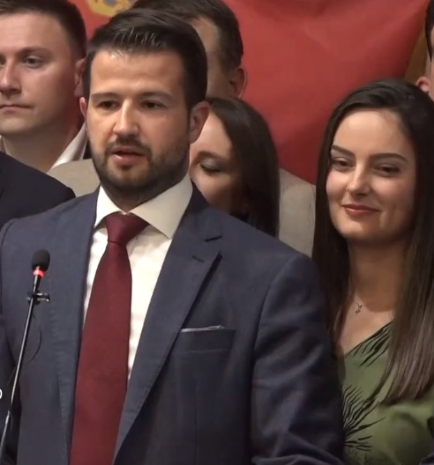 Ein Mann im Anzug steht an einem Rednerpult und spricht in ein Mikrofon, mit einem Banner im Hintergrund und einer Menge davor.