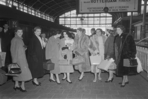 Schwarzes und weißes Bild von Frauen, die an einem Bahnhof stehen und Taschen halten, mit einem Zug auf der linken Seite und Geländern und einer Tafel auf der rechten Seite.
