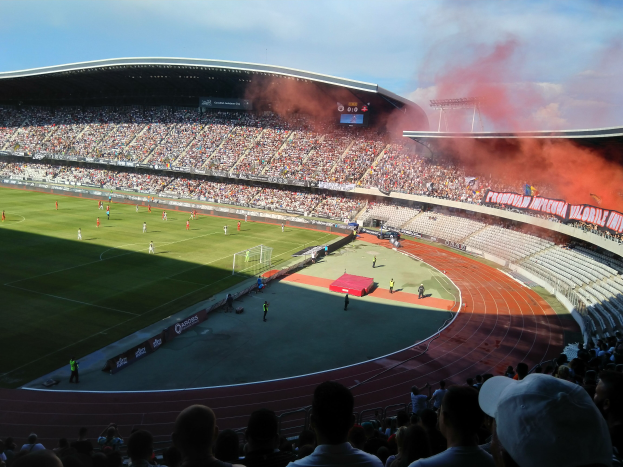 Ein großes Stadion voller Zuschauer, die ein Fußballspiel verfolgen, mit Spielern im Einsatz auf dem Feld und Text am unteren Bildrand.