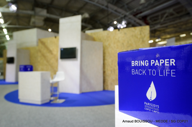 Ein Messestand mit einem blauen und weißen Schild, auf dem "Bring Paper Back to Life" steht, auf der rechten Seite, mit unscharfen Stühlen, Tischen, einem Wandfernseher und Deckenleuchten im Hintergrund.